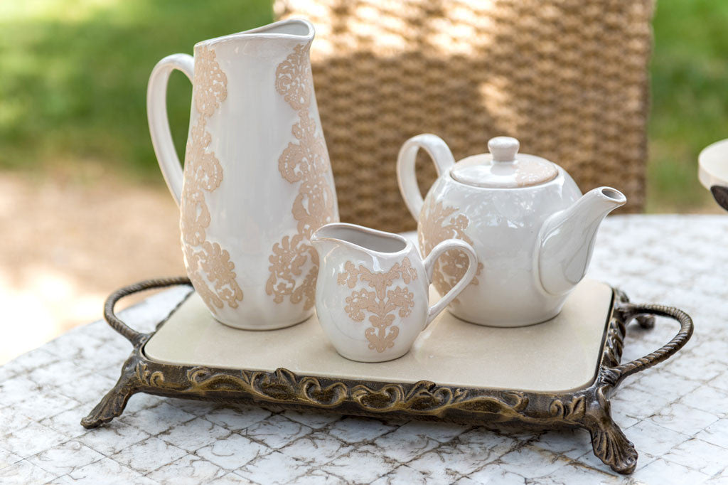 White ceramic vessel with blush on white, lace-inspired embossed design shown on a water pitcher, in a scene with other tea party serveware at an outdoor picnic