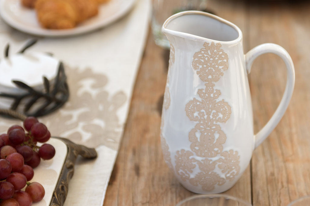 White ceramic vessel with blush on white, lace-inspired embossed design shown on a water pitcher in a table scene with grapes on a platter
