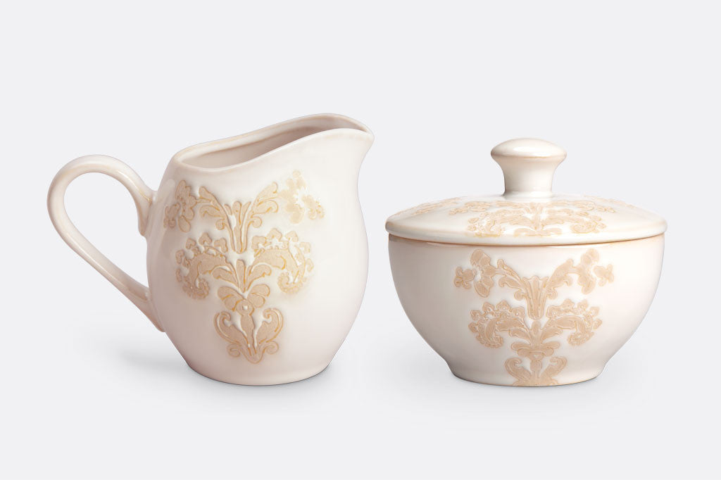 White ceramic vessel with blush on white, lace-inspired embossed design shown on cream and sugar set