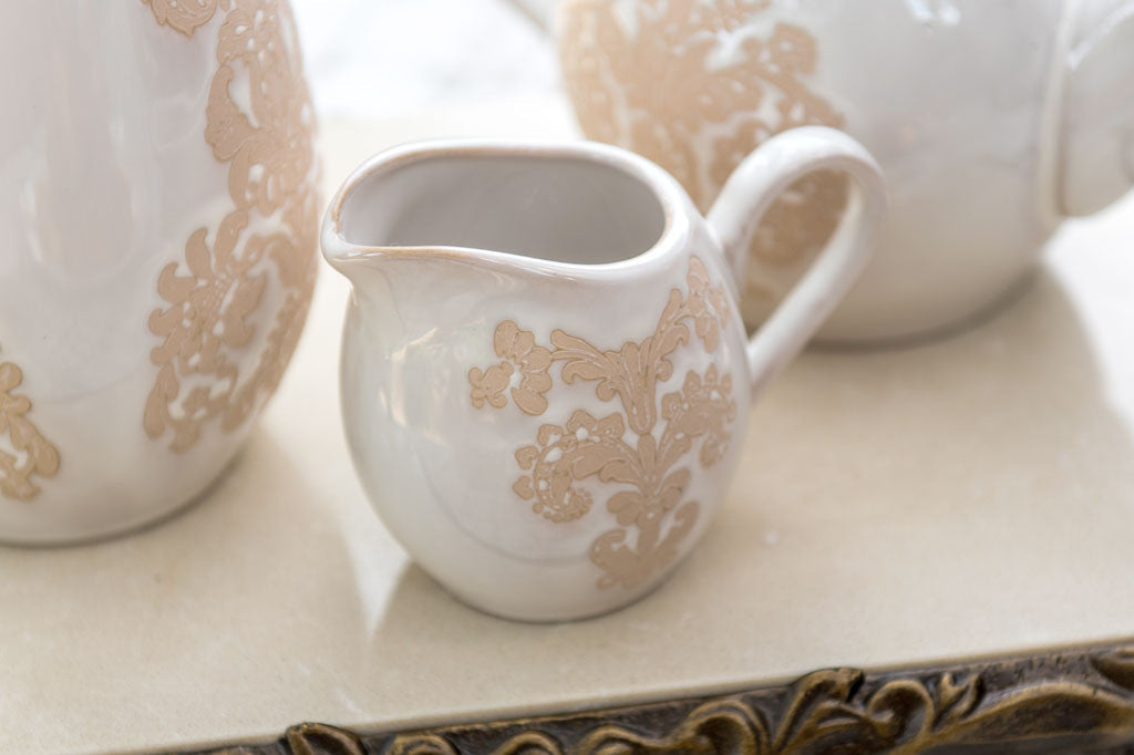 White ceramic vessel with blush on white, lace-inspired embossed design shown on cream and sugar set - close up view of pattern on the creamer