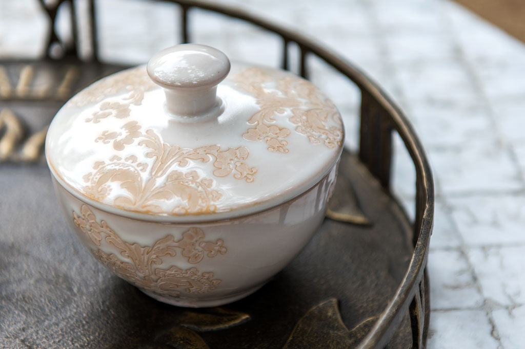 White ceramic vessel with blush on white, lace-inspired embossed design shown on cream and sugar set, closeup of lid upon the sugar bowl