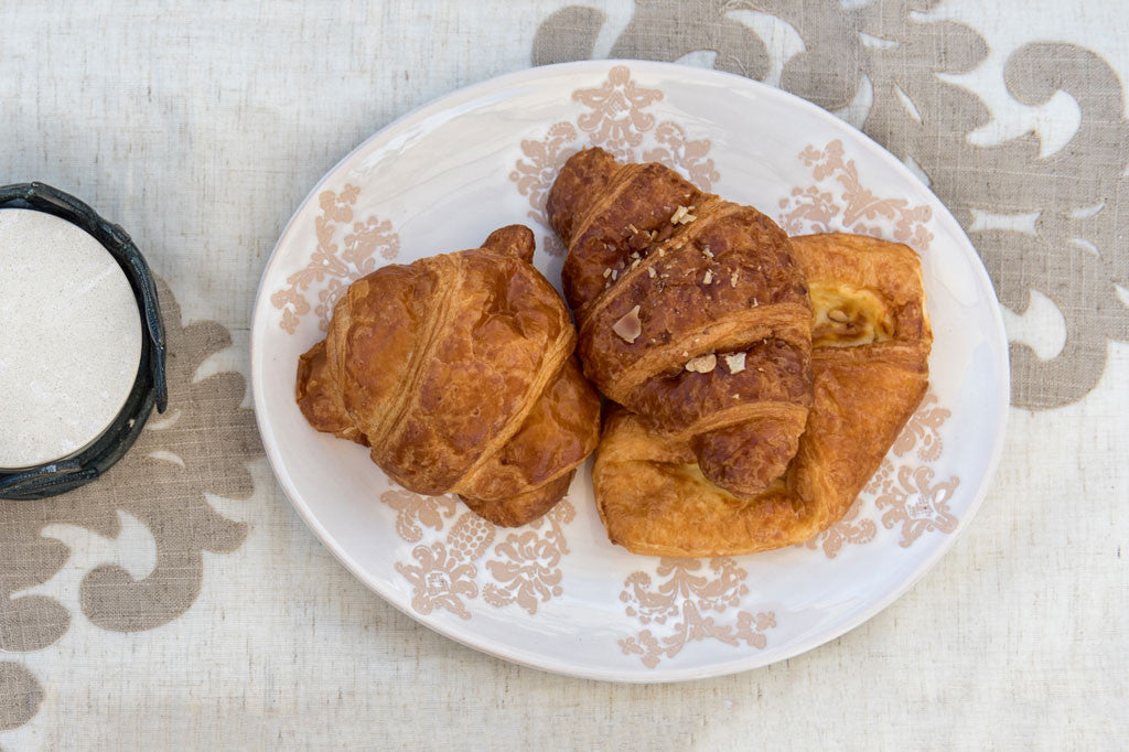 White ceramic vessel with blush on white, lace-inspired embossed design shown on an oval serving plate holding 3 croissants on a table, top view