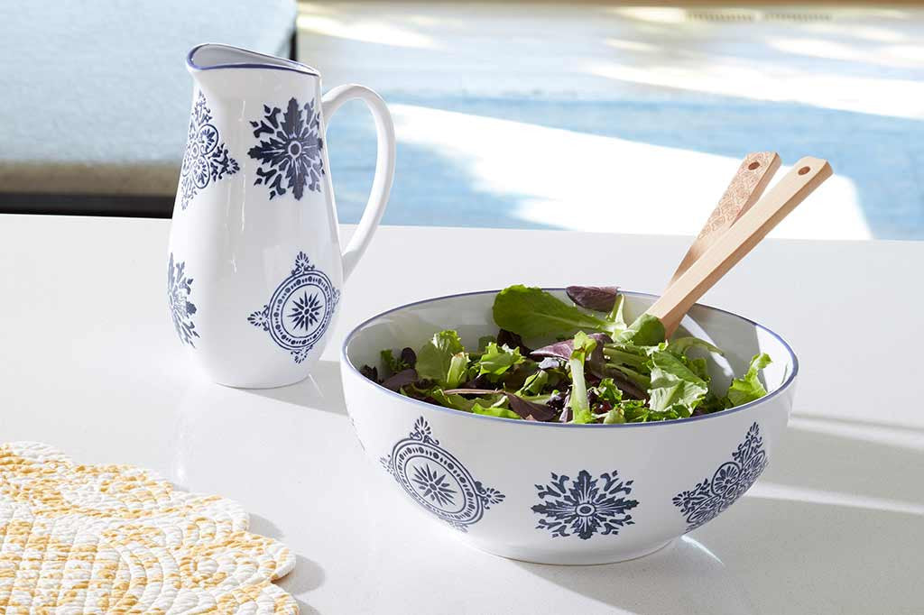 white ceramic bowl with blue mediterranean glazed print shown on table with pitcher; it holds salad and wood spoons