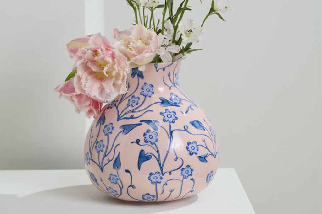 Ceramic vase with base color of pink and a vine and blossom blue floral - shown with pink flowers on a table