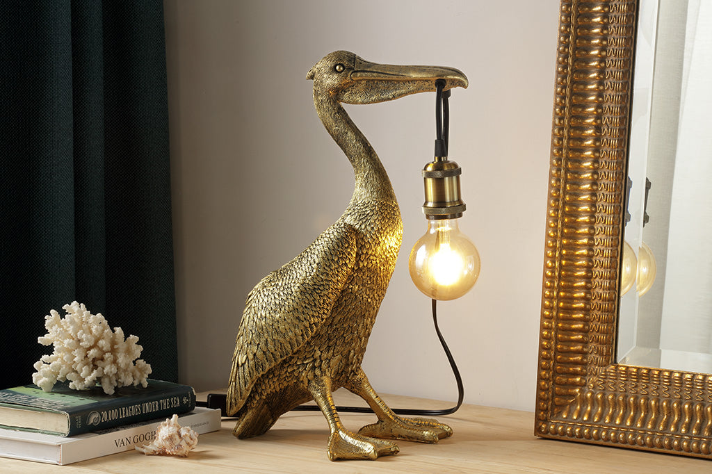 novelty desk lamp of pelican holding lightbulb by the beak - gold finished resin, shown on desk by shells and a mirror 