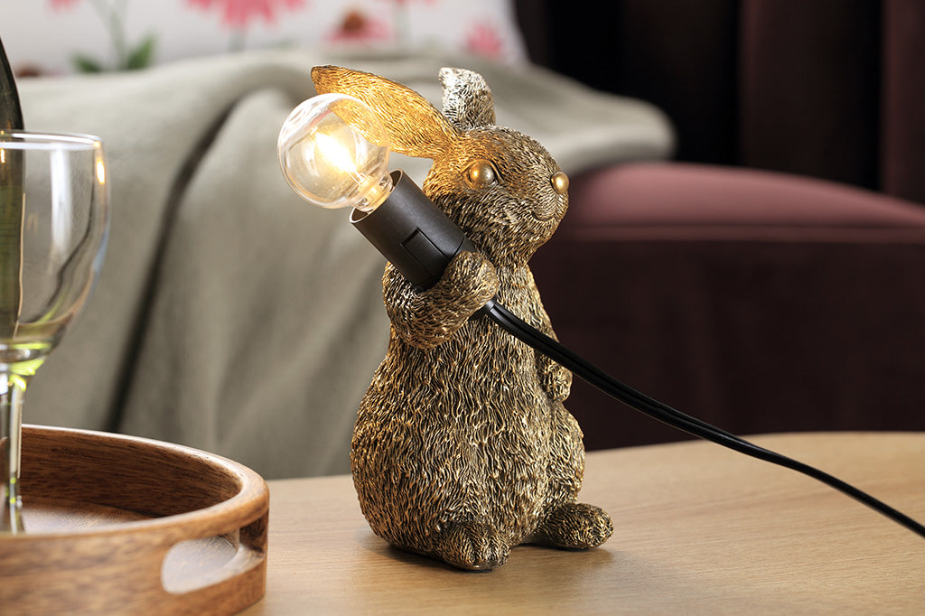 novelty desk lamp of gold finished resin. A rabbit holding lightbulb over its shoulder shown in livingroom setting on coffee table