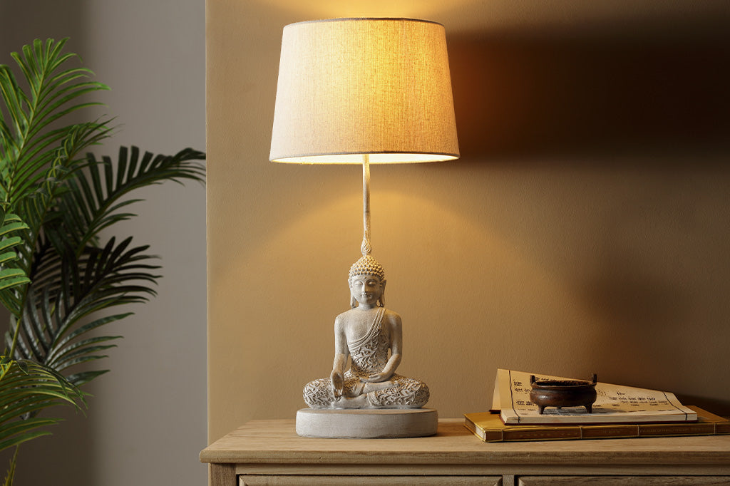 cream colored Resin Buddha sculpture table lamp with cream fabric shade shown on desk near houseplant