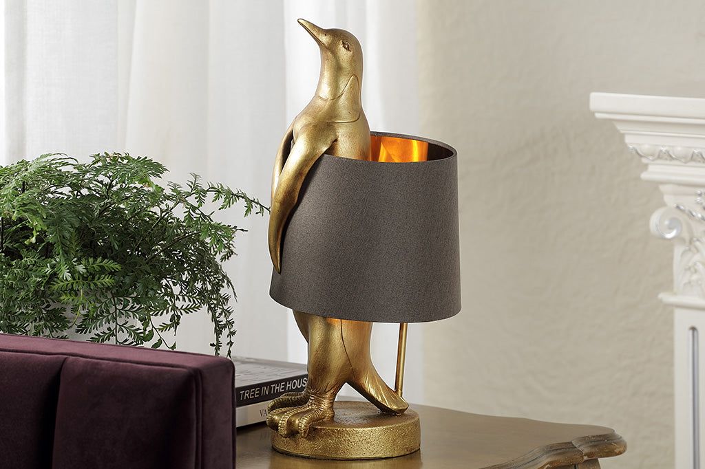 novelty table lamp of gold finished resin penguin wearing a brown fabric lampshade around its torso. Shown on console table in livingroom setting by houseplant