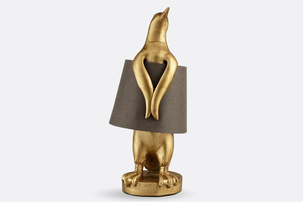 whimsical table lamp of gold finished resin penguin wearing a brown fabric lampshade around its torso.