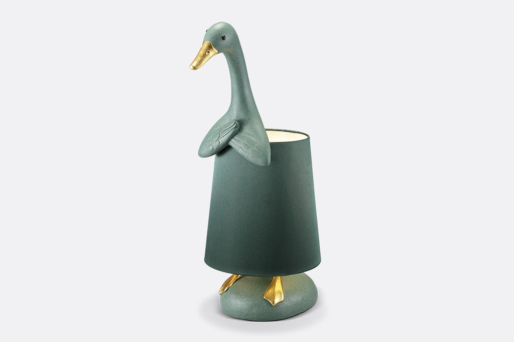 face left view of whimsical table lamp, resin. Green duck wearing a lampshade around its torso with gold webbed feet sticking out on lamp base.