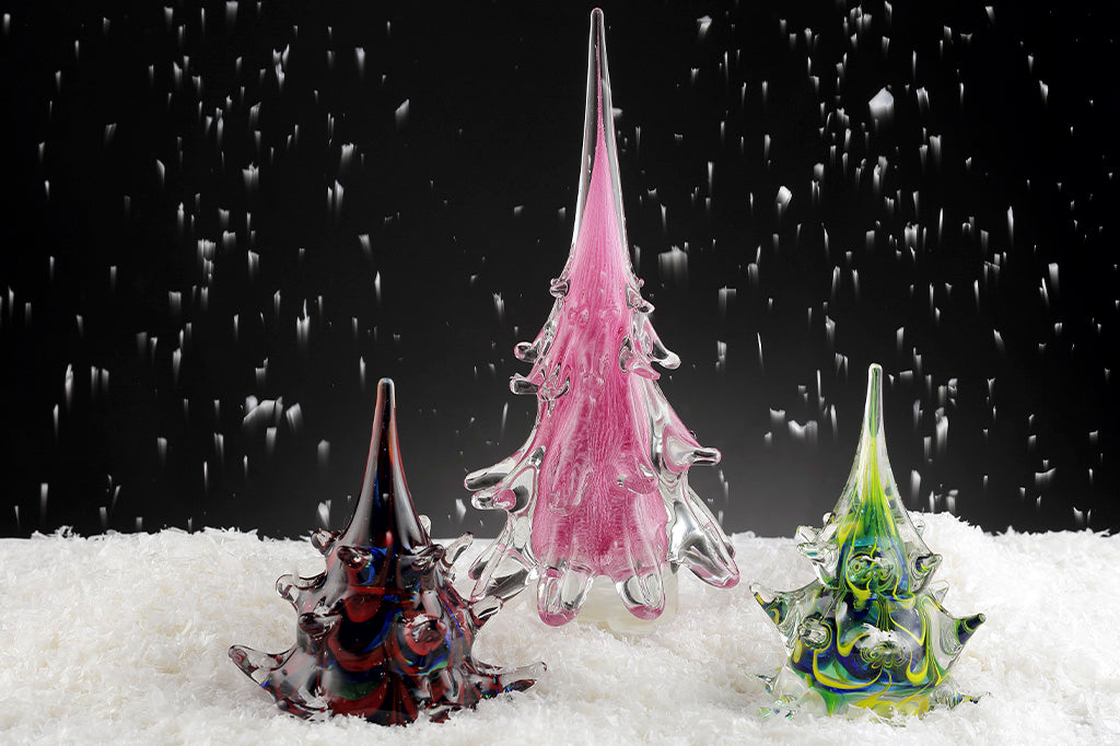 artificial snown falling on scene of holiday table scene of three glass christmas trees in lavender, green/blue, and red/blue