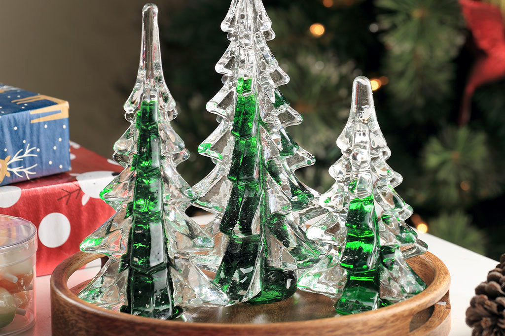 Close up view of Set of 3 glass green and clear trees, used as a holiday decoration on a wood tray surrounded by presents; Christmas tree in background