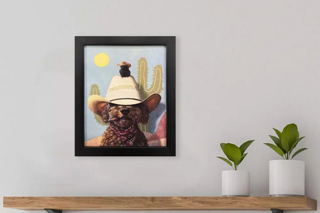 Framed artwork of a dog wearing a cowboy hat and a bird wearing a cowboy hat on top of him with a cactus and sun in the background.