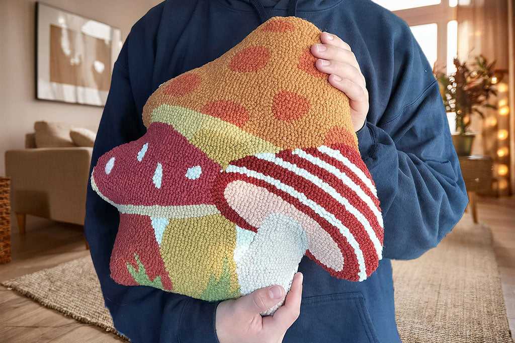 Hooked wool throw pillow featuring 3 mushrooms in red and orange hues, held by person in black hoodie in sunny livingroom with beige couch and rug