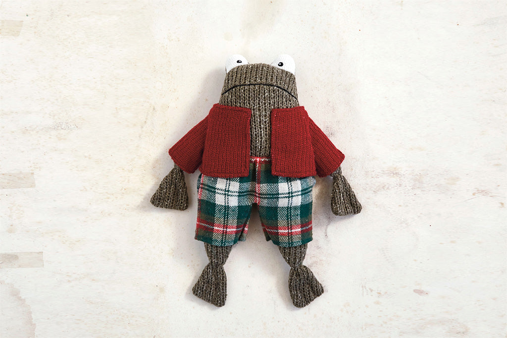 Knitted stuffed frog wearing red sweater and plaid pants shown on table surface