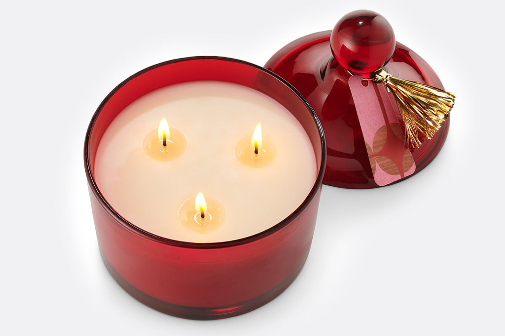 Red glass lidded candle shown with 3 lit wicks and gold tassle tag