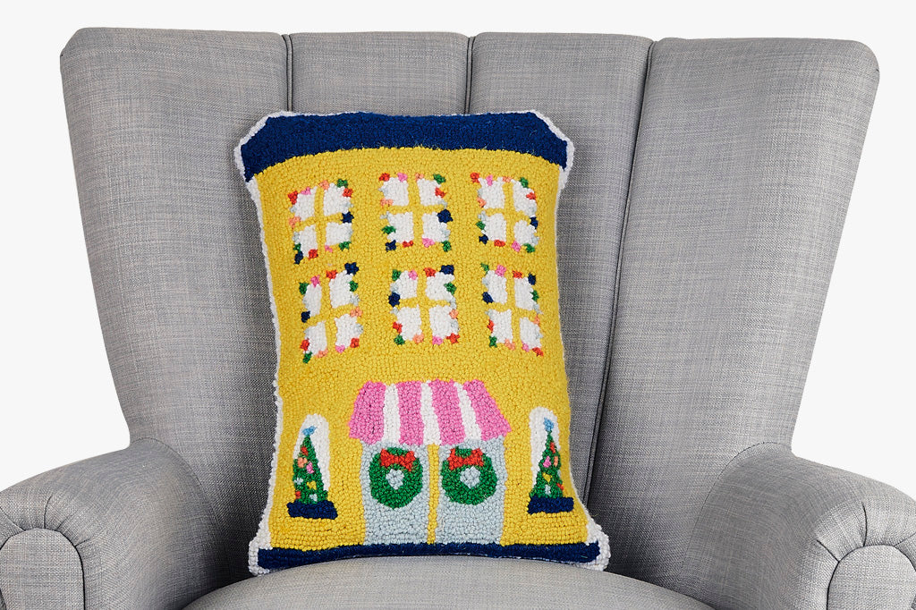 hooked wool pillow features a charming, shaped yellow hotel adorned with festive holiday lights- shown on grey loveseat