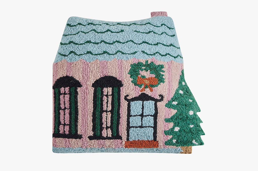 Hooked wool pillow in pink house shape adorned with a blue roof, double windows, and a festive wreath and Christmas tree