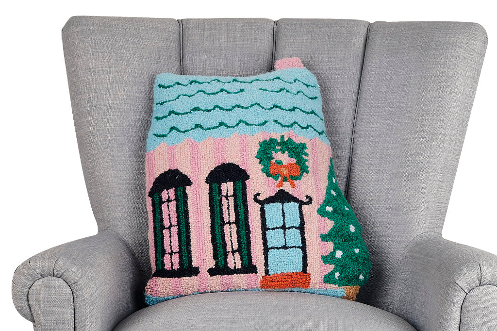 Hooked wool pillow in pink house shape adorned with a blue roof, double windows, and a festive wreath and Christmas tree shown on grey couch