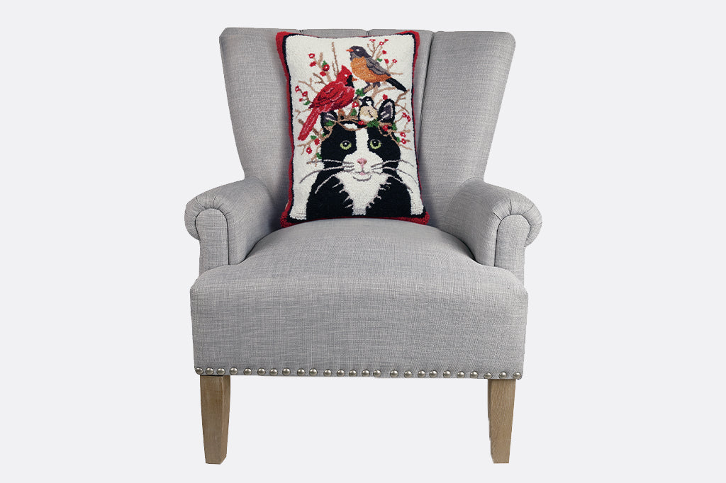 Black cat with pair of birds seated on berry branches hooked pillow, seated on gray arm chair 