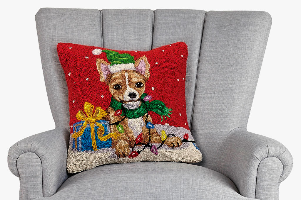 Hooked wool pillow with design of chihuahua, dressed in a green hat and scarf. Pillow displayed on grey loveseat.