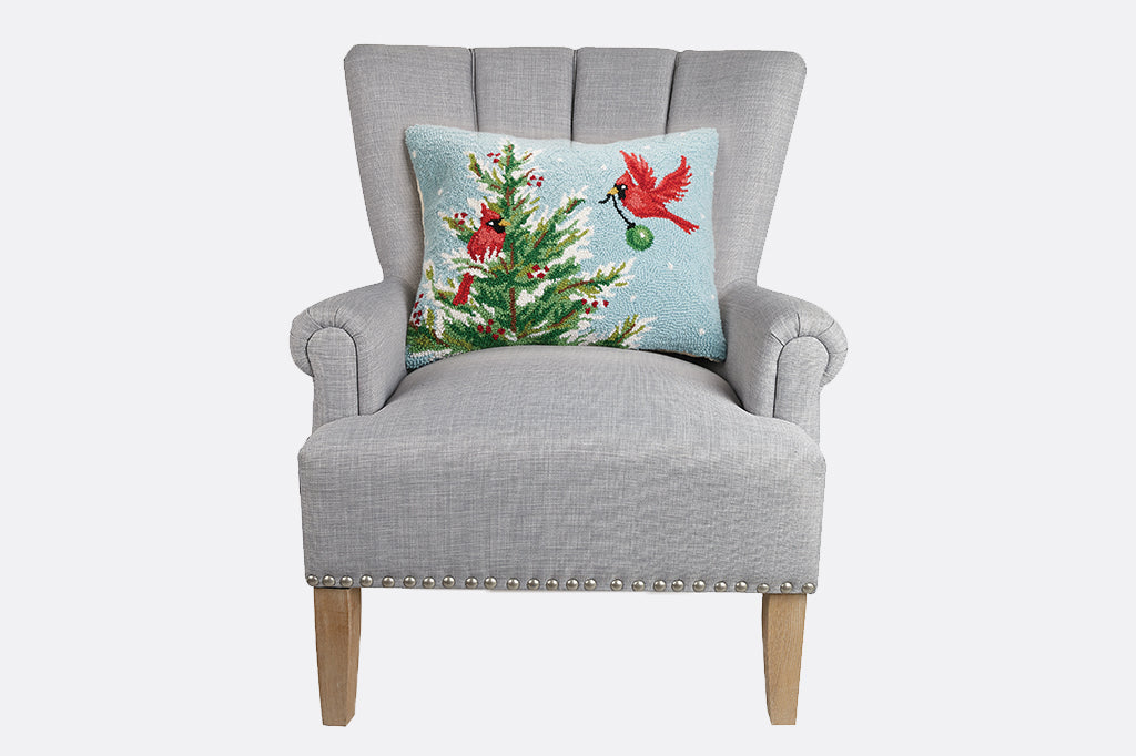 holiday tree being decorated by two cardinal hooked pillow seated on gray armchair 