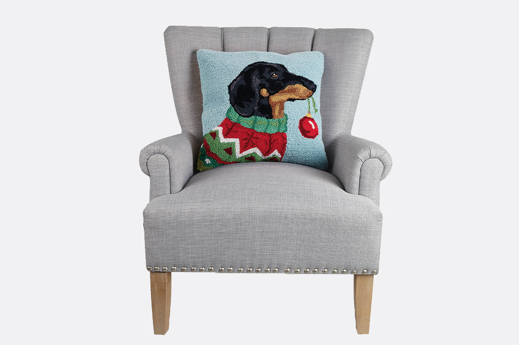 Hooked dog holding ornament in mouth wearing a sweater seated on gray arm chair 