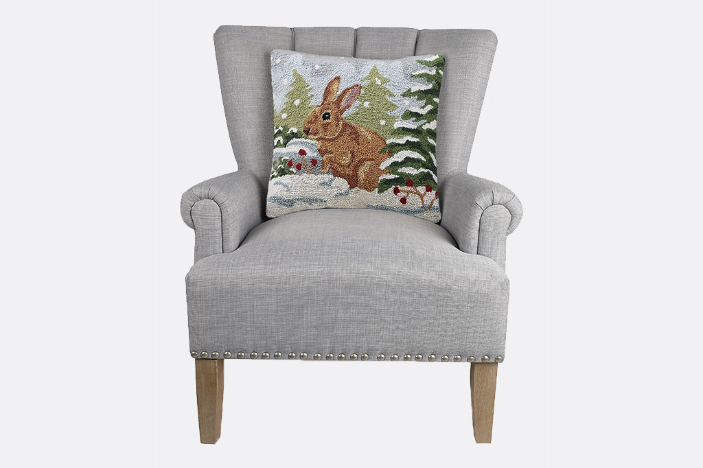 brown bunny in snowy forest scene hooked pillow. set on gray arm chair 