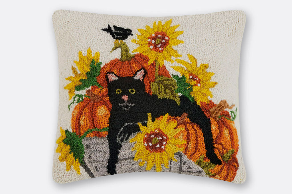 Hooked wool pillow shows black cat lounging on a wagon of pumpkins and sunflowers on a cream background; black crow atop stem