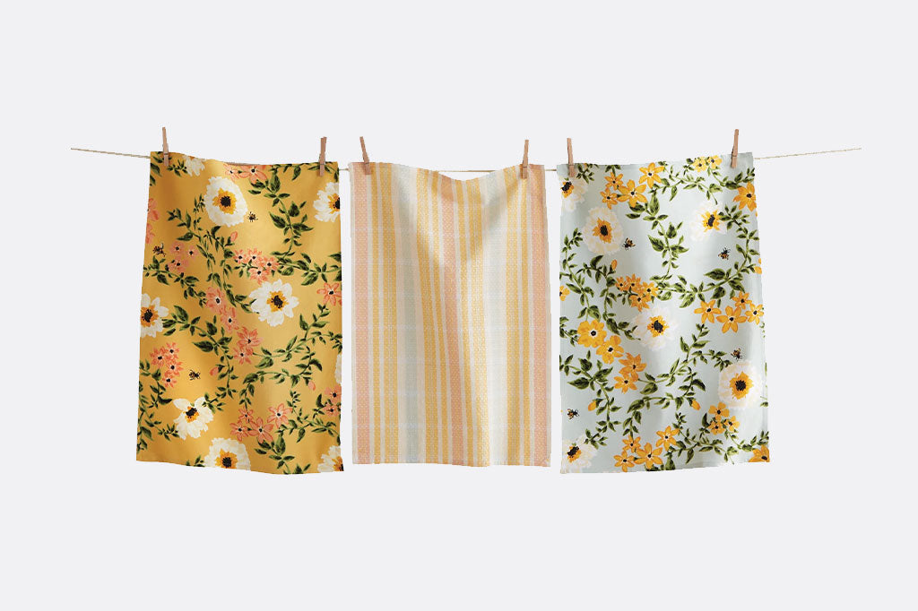 3 dishtowels hung on a clothesline; all have bright, cheerful prints in yellow. Two floral and 1 plaid