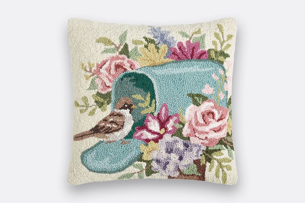 hooked wool throw pillow showing blue mailbox surrounded by flowers. bird rests on open mailbox door. cream background