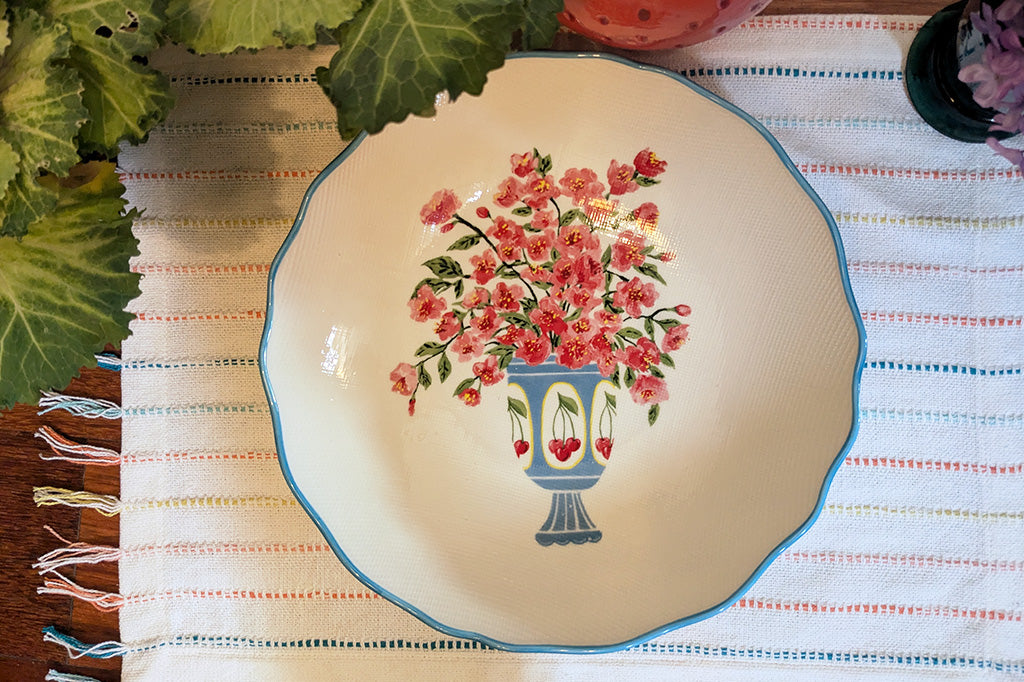 Stoneware bowl with scalloped edges painted in blue with floral arrangement in cheery vase . Bowl shown on placement with colorful stripes