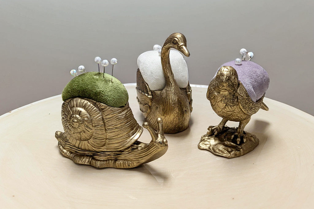 Three decorative pin cushions shaped like a snail, swan, and goose on a neutral background.