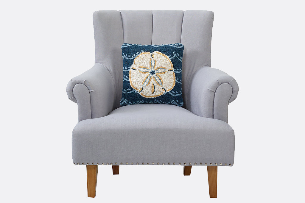 Hooked pillow with cream colored sand dollar shell on dark blue ocean background with wave design. Propped up on a gray arm chair 