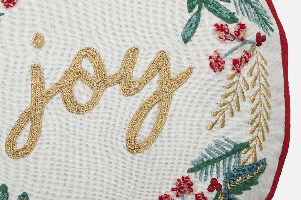 Detail on throw pillow of embroidered word 'joy' with floral decorations on a white background
