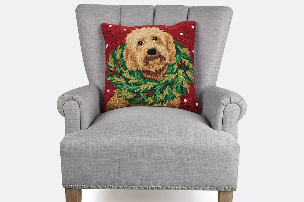 Hooked wool throw pillow with design on brown labradoodle wearing a holiday wreath on its neck; shown on a grey armchair