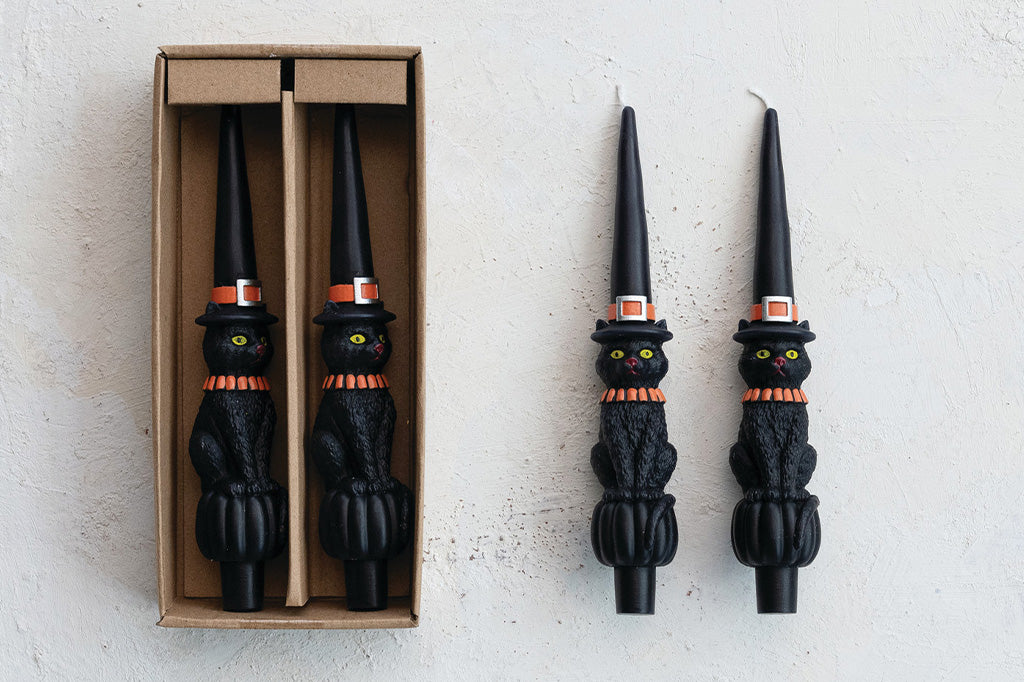 Black cat-shaped candles with orange collars and green eyes, one in a box on a light gray background.