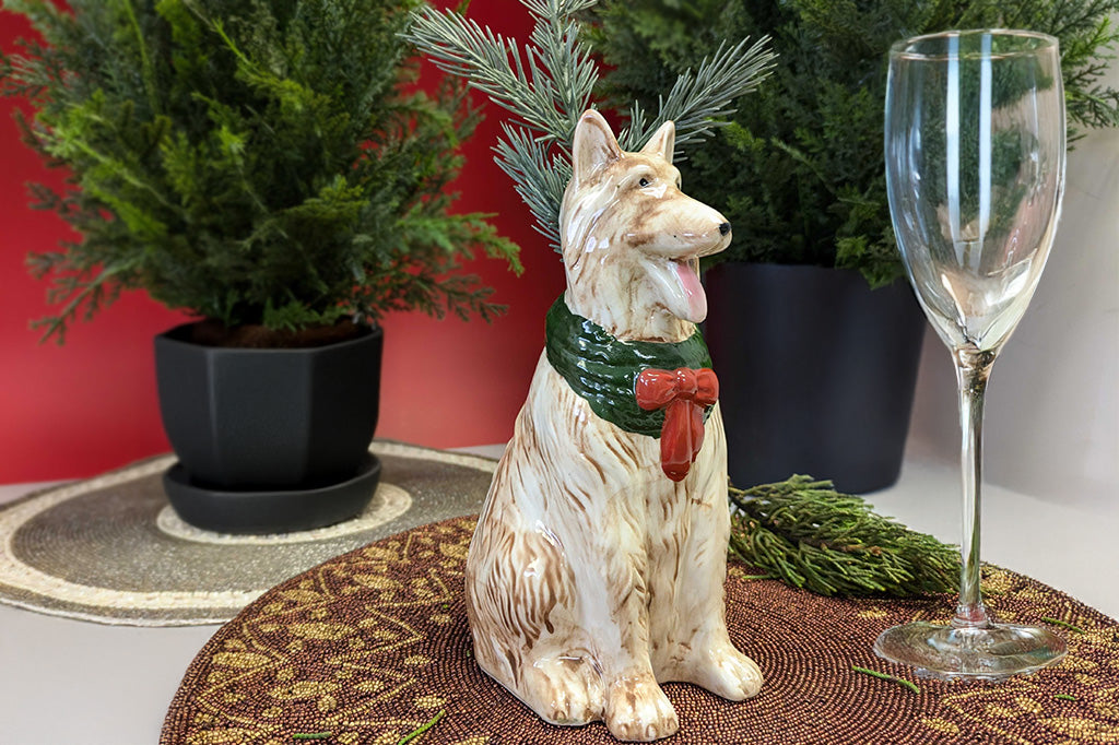 Dog bud vase with a green scarf and red bow tie on a table with a wine glass and plants.