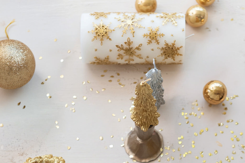 Decorative tree candles and a white pillar candle that has gold snowflakes - all shown on white table with Christmas ball ornaments and gold glitter