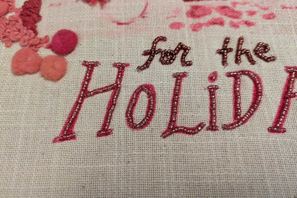Embroidered text 'for the Holidays' on fabric with holiday decorations.