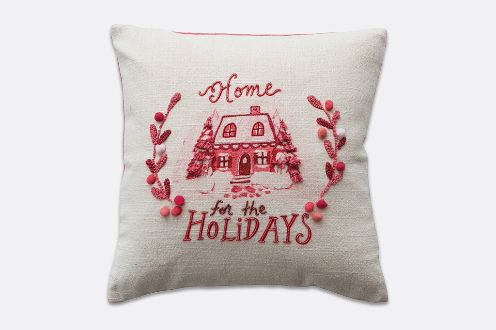 A white pillow with pink and red accents, the imagery of a snowed in house and the words "home for the holidays" embroidered onto it.