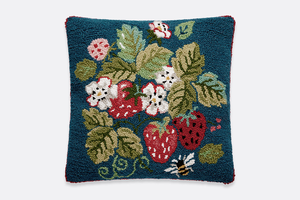 A hooked pillow with illustrations of strawberries, flowers, leaves, and a bee.