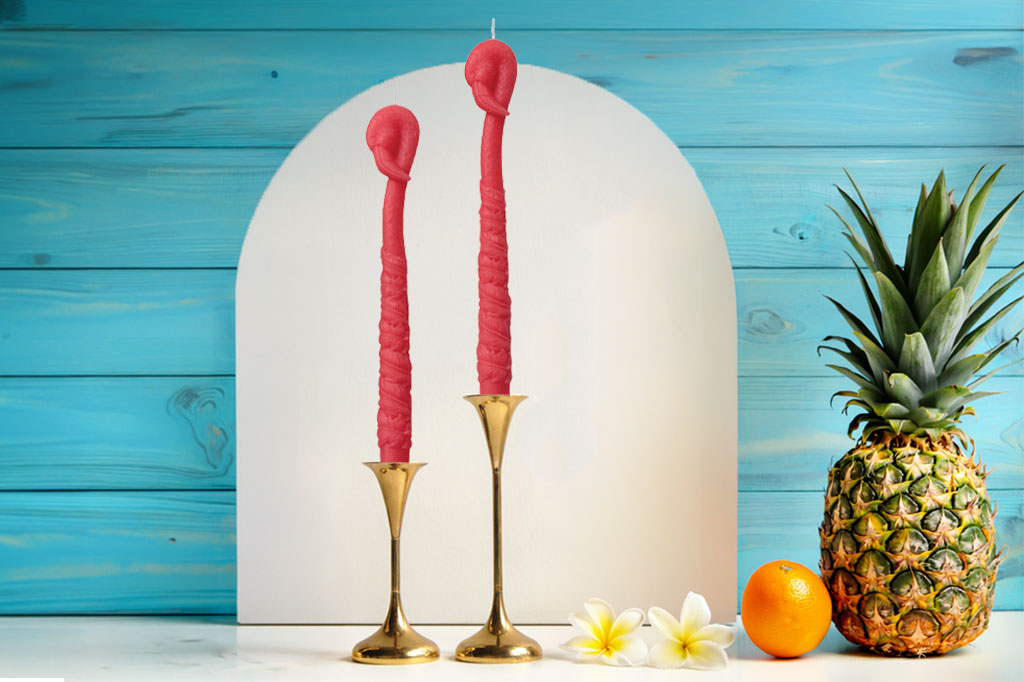 Two pink flamingo candles in gold holders with a pineapple and orange in front of a blue wooden background