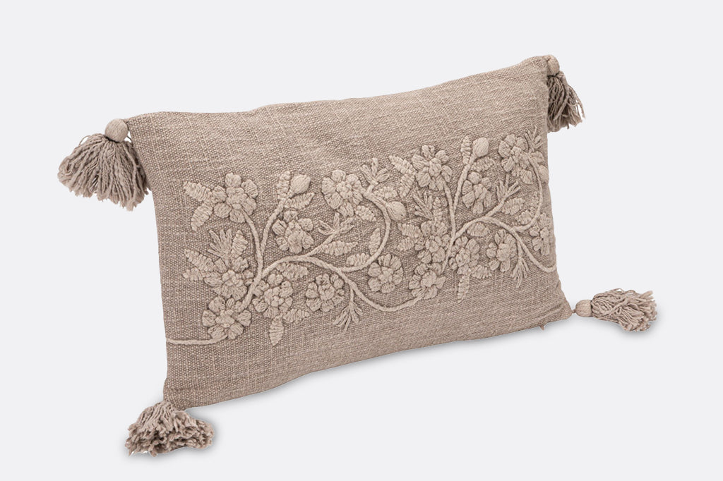 A beige, tassel pillow with embroidered details of flowers and leaves.