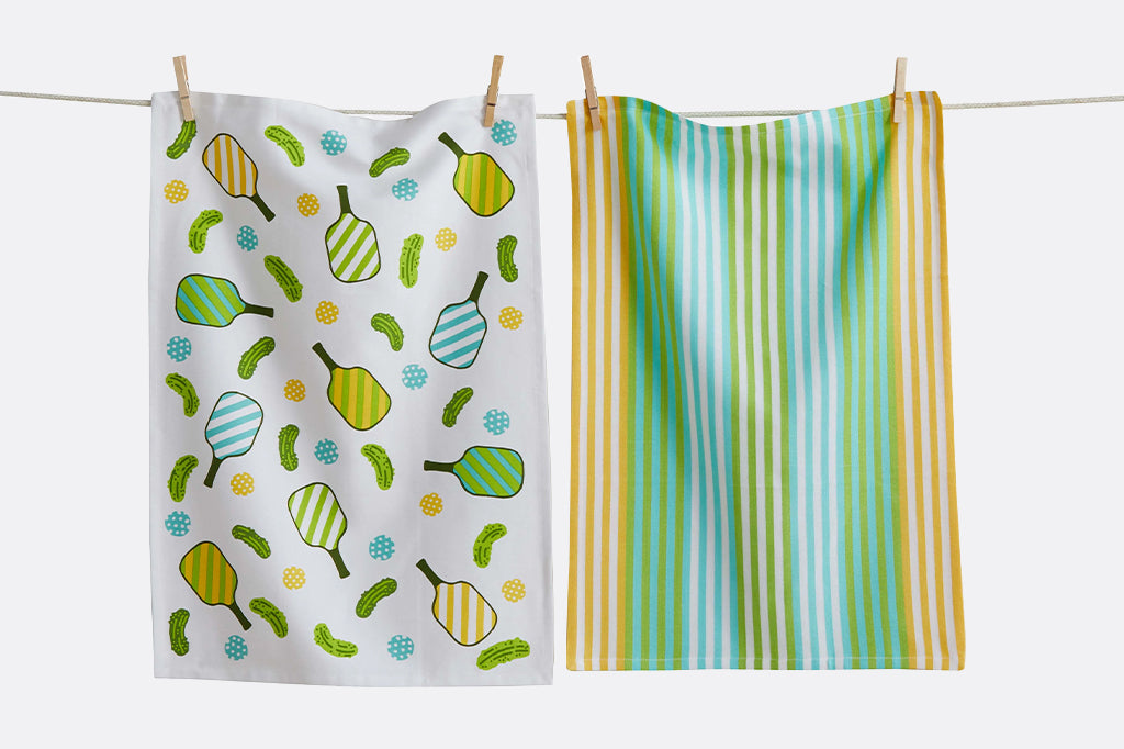 A set of dishtowels with illustrations of pickles, pickleballs, paddles, and stripes.