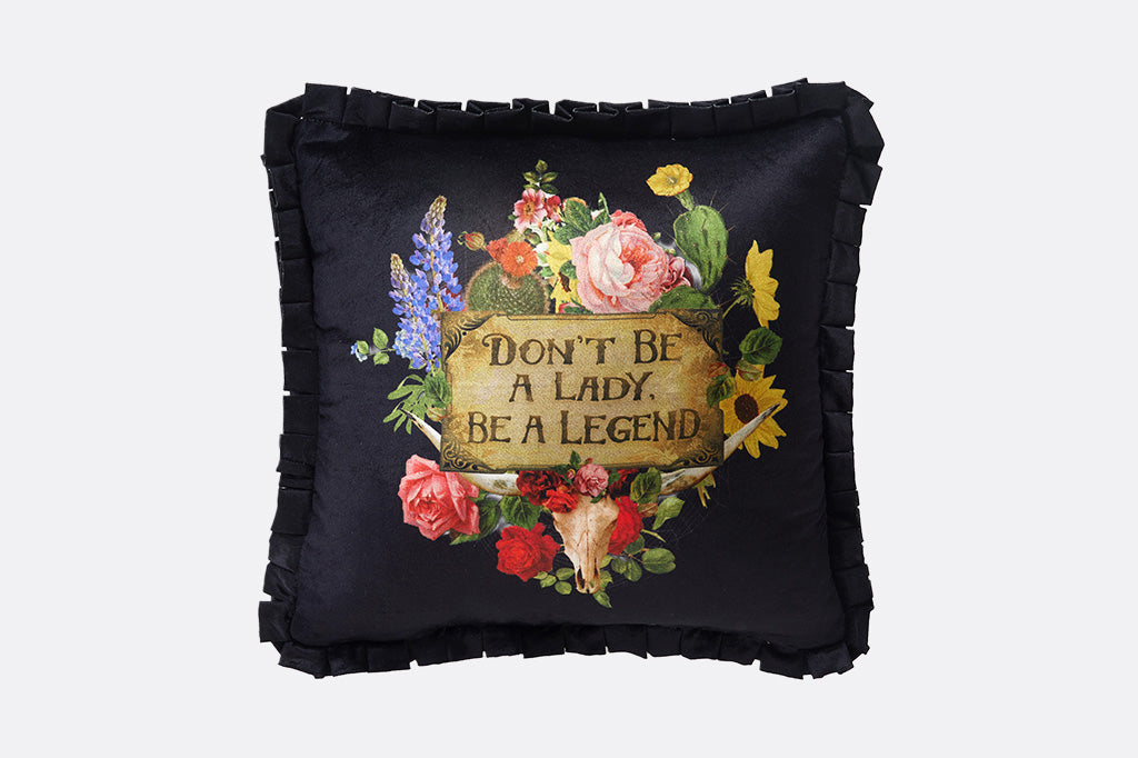 A black pillow with flower and cacti, with a sign that says "Don't be a lady, be a legend."