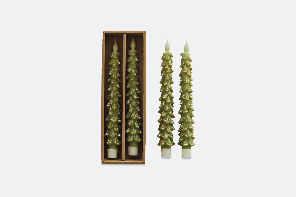 Two flameless candles, shaped like christmas trees with green coloring and gold highlights, pictured in packaging next to two more. 