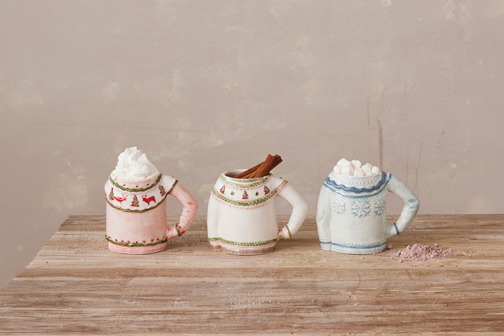Three small ceramic mug-shaped ornaments or figurines displayed side by side on a wooden table. The left one is pale pink with a red and green holiday sweater pattern; the middle one is white with a light blue knit pattern; the right one is white with a beige knit pattern.