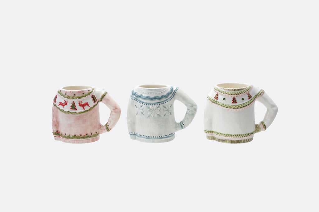 Three small ceramic mug-shaped ornaments or figurines displayed side by side on a light gray background. The left one is pale pink with a red and green holiday sweater pattern; the middle one is white with a light blue knit pattern; the right one is white with a beige knit pattern.
