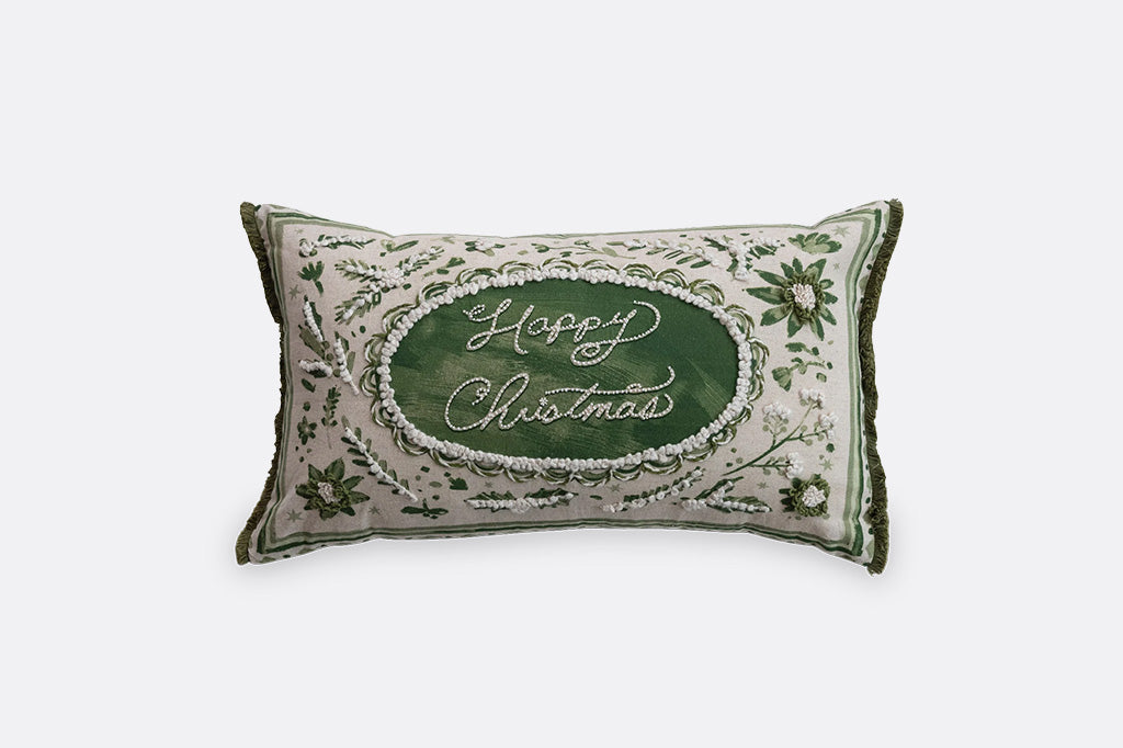 A rectangular Christmas throw pillow with a cream-colored embroidered background, a central dark green oval containing the words 'Happy Christmas' in white script, and green floral and vine motifs around the border.